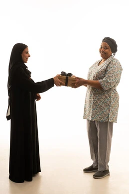 النظر للكاميرا اثناء تلقى الهدية ,بورتريه خلفية بيضاء الجسم بالطول الكامل لعاملة منزلية افريقية سمراء البشرة ترتدي يونيفورم باللون الرمادي تستقبل الهدايا من امرأة عربية خليجية سعودية محجبة ترتدي عباءة سوداء