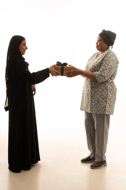 ايماءات سعادة وفرحة على وجه امراة عربية خليجية سعودية محجبة ترتدي عباءة سوداء اثناء تقديم هدية لعاملة منزلية افريقية سمراء البشرة ترتدي يونيفورم باللون الرمادي ,بورتريه خلفية بيضاء الجسم بالطول الكامل