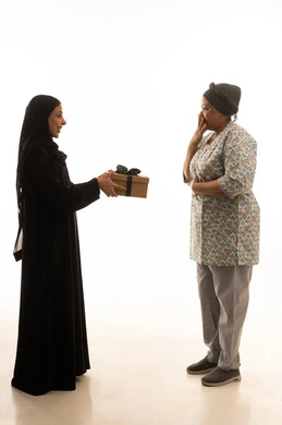 Expressions of surprise and joy on the face of a dark-skinned African domestic worker wearing a gray uniform, a portrait with a white background of a Saudi Arabian Gulf woman in a hijab wearing a black abaya while presenting a gift to her maid, full-body.