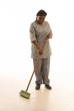 عاملة منزلية افريقية ملونة سمراء البشرة ترتدي يونيفورم لون رمادي تقوم بكنس الارضية بسعادة و سرور , بورتريه خلفية بيضاءالجسم بالطول الكامل