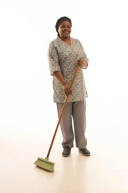 السعادة اثناء مزاولة اعمال النظافة ,بورتريه خلفية بيضاء لعاملة منزلية افريقية سمراء البشرة ترتدي يونيفورم اللون رمادي تحمل مكنسة يدوية ,النظر للكاميرا مع الابتسام ,الجسم بالطول الكامل