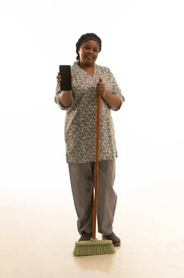 استخدام الهاتف المحمول والتقنية الحديثة في اعمال النظافة ,عاملة منزلية افريقية داكنة البشرة ترتدي يونيفورم باللون الرمادي تحمل مكنسة يدوية , عرض شاشة هاتف سوداء فارغة ,الجسم بالطول الكامل ,النظر للكاميرا