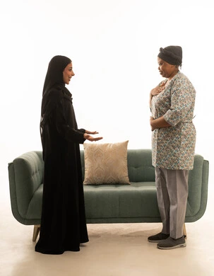 بورتريه خلفية بيضاء الجسم بالطول الكامل ,امرأة عربية خليجية سعودية محجبة ترتدي عباءة سوداء و عاملة منزلية افريقية سمراء البشرة ترتدي يونيفورم باللون الرمادي تجريان حديثًا شيقًا في غرفة المعيشة ,اريكة باللون الرمادي