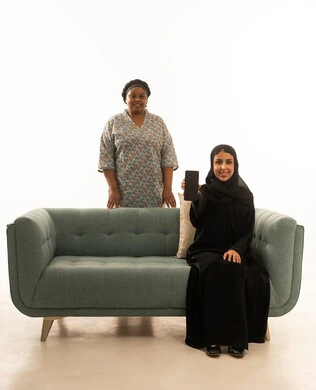 Sitting happily on a gray sofa, a portrait with a white background featuring a smiling Saudi Arabian Gulf woman in a hijab wearing a black abaya and an African housemaid with dark skin wearing a gray uniform, displaying a blank black mobile phone screen, smiling happily at the camera.