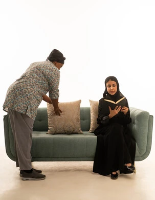 عاملة منزلية افريقية سمراء البشرة ترتدي يونيفورم باللون الرمادي اثناء متابعة الاعمال الروتينية للعناية بالمنزل ,بورتريه خلفية بيضاء لامرأة عربية خليجية سعودية محجبة ترتدي عباءة سوداء تجلس على اريكة رمادية اللون اثناء مطالعة كتاب ,الجسم بالطول الكامل