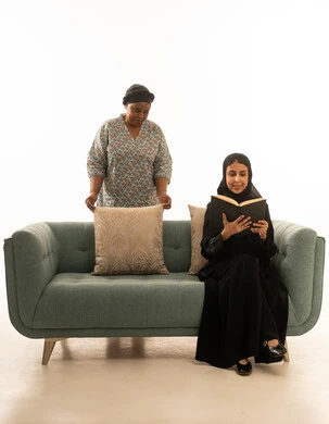 مطالعة كتاب اثناء متابعة عملية تنظيف المنزل ,امرأة عربية خليجية سعودية محجبة ترتدي عباءة سوداء تجلس على اريكة رمادية اللون ,بورتريه خلفية بيضاء لعاملة منزلية افريقية داكنة البشرة ترتدي يونيفورم باللون الرمادي تقوم بترتيب الوسائد