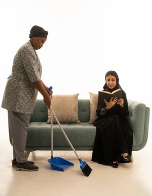 امرأة عربية خليجية سعودية محجبة تقرأكتاب اثناء الجلوس على اريكة باللون الرمادي ,بورتريه خلفية بيضاء الجسم بالطول الكامل لعاملة منزلية افريقية سمراء البشرة ترتدي يونيفورم باللون الرمادي تنظف ارضية غرفة المعيشة باستخدام مكنسة يدوية زرقاء اللون