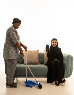 A portrait with a white background of a Saudi Arabian Gulf woman wearing a hijab, smiling at the camera while displaying a blank black mobile phone screen. The full-body image features a dark-skinned African domestic worker wearing a gray uniform, sweeping the floor with a hand broom.