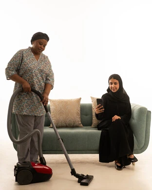 تصفح الهاتف المحمول اثناء الجلوس على أريكة غرفة المعيشة رمادية اللون ,سيد ة عربية خليجية سعودية مبتسمة للهاتف ترتدي عباءة سمراء ,بورتريه خلفية بيضاء الجسم بالطول الكامل لشغالة افريقية ملونة سمراء البشرة ترتدي يونيفورم باللون الرمادي