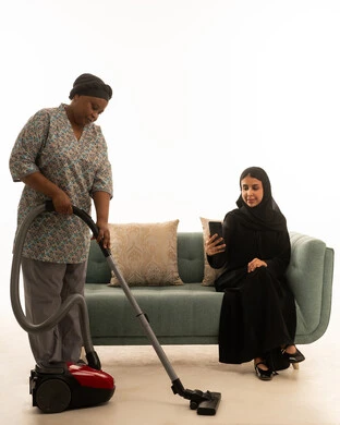 Using a mobile phone while following up on daily household chores, an Arab Gulf Saudi woman in a hijab wearing a black abaya sitting on a gray sofa, a portrait with a white background of an African housekeeper with dark skin wearing a gray uniform vacuuming the floor with a vacuum cleaner, full body.