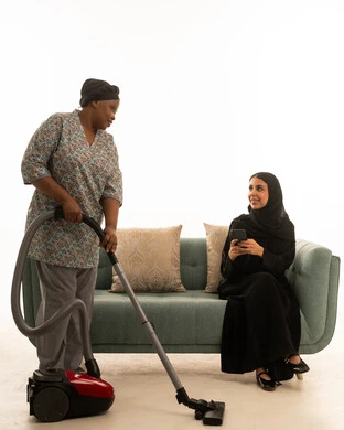الاستعانة بالاجهزة الحديثة في العناية بالمنزل ,امراة عربية خليجيةسعودية محجبة ترتدي عباءة سوداء تستخدم الهاتف المحمول اثناء متابعة العاملة المنزلية ,شغالة افريقية سمراء البشرة ترتدي يونيفورم باللون الرمادي تستخدم المكنسة الكهربائية ,خلفية بيضاء الجسم بالطول الكامل