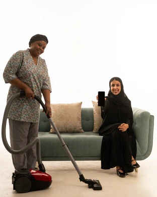 التطور في اداء الاعمال المنزلية باستخدام الاجهزة الكهربائية الحديثة , امرأة عربية خليجية سعودية ترتدي عباءة سوداء ,عرض شاشة هاتف محمول فارغة ,بورتريه خلفية بيضاء الجسم بالطول الكامل لعاملة منزلية افريقية سمراء البشرة تكنس الارض بالمكنسة الكهربائية , النظر للكاميرا