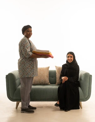 متابعة الاعمال المنزلية اليومية ,امرأة عربية خليجية سعودية محجبة ترتدي عباءة سوداء تجلس على اريكة رمادية اللون ,بورتريه خلفية بيضاء الجسم بالطول الكامل لعاملة منزلية سمراء البشرة مبتسمة ترتدي يونيفورم باللون الرمادي , النظر للكاميرا