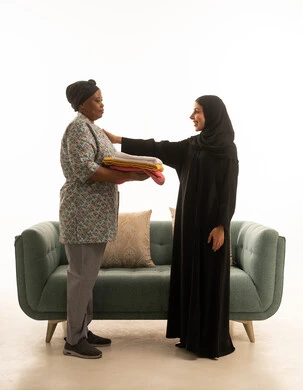 ربة منزل عربية خليجية سعودية محجبة ترتدي عباءة سوداء تربت على كتف شغالة افريقية و تثني على عملها ,بورتريه خلفية بيضاء الجسم بالطول الكامل لعاملة منزلية سمراء البشرة ترتدي يونيفورم باللون الرمادي تقف امام الاريكة باللون الرمادي ,