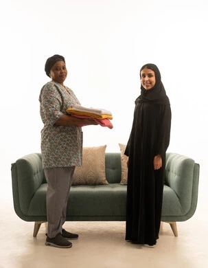 Full-length body portrait, white background, a Saudi Arabian Gulf woman in a hijab wearing a black abaya smiling at the camera, an African housemaid with dark skin wearing a gray uniform holding towels, standing in front of a gray sofa.