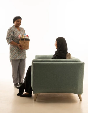 An African domestic worker with dark skin wearing a gray uniform carrying a wicker basket filled with household cleaning supplies, a portrait with a white background of a Saudi Arabian Gulf woman wearing a black abaya sitting on a gray sofa.
