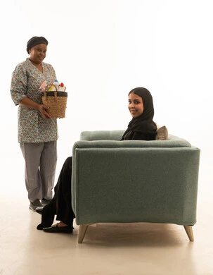 Looking at the camera with a smile, an Arab Gulf Saudi woman wearing a black abaya and a hijab, an African domestic worker with brown skin wearing a gray uniform holding a wicker basket full of cleaning supplies, full-length portrait with a white background.