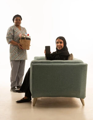 شراء مستلزمات العناية بالمنزل باستخدام الهاتف المحمول ,امرأة عربية خليجية سعودية محجبة ترتدي عباءة سوداء تعرض شاشة جوال فارغة سوداء للكاميرا ,عاملة منزلية افريقية سمراء البشرة ترتدي يونيفورم باللون الرمادي تحمل المنظفات داخل سلة قش ,بورتريه خلفية بيضاء الجسم بالطول الكامل