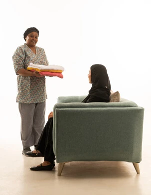 ربة منزل عربية خليجية سعودية محجبة ترتدي عباءة سوداء تجلس على اريكة رمادية ,عاملة منزلية افريقية ذات بشرة سمراء ترتدي يونيفورم لون رمادي تنظر للكاميرا بابتسامة , بورتريه خلفية بيضاء بالطول الكامل للجسم ,