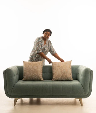 الوقوف بشكل مائل امام اريكة رمادية اللون ,بورتريه خلفية بيضاء لعاملة منزلية افريقية سمراء البشرة ترتدي يونيفورم لون رمادي وغطاء رأس ,الطول الكامل للجسم