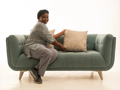 النظر للكاميرا بابتسامة وسعادة ,عاملة منزلية سمراء البشرة ترتدي يونيفورم لون رمادي ترتب وسادات اريكة رمادية ,بورتريه خلفية بيضاء