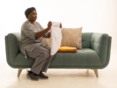 شغالة منزلية افريقية ذات بشرة داكنة ترتدي يونيفورم لون رمادي تقوم بطي منشفة ,اريكة رمادية ,بورتريه خلفية بيضاء