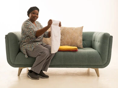 العمالة المرخصة المؤهلة لادارة المنزل .بورتريه لعاملة منزلية ملونة سمراء البشرة ترتدي يونيفورم رمادي اللون  تجلس على أريكة , ترتيب وتصفيط الملابس بسعادة ,خلفية بيضاء