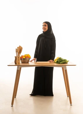 امرأة عربية خليجية سعودية محجبة ترتدي عباءة سوداء بجانب طاولة تحضير طعام خشبية , بورتريه خلفية بيضاء الجسم بالطول الكامل ,النظر للاعلى الى شئ ما