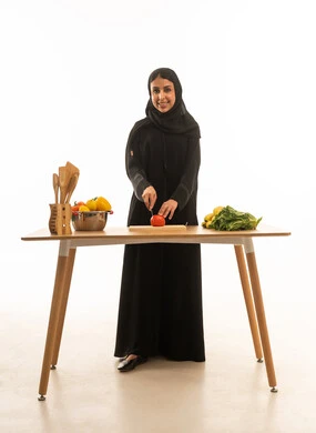 Portrait of a veiled Saudi Arabian woman from the Gulf region, wearing a black abaya, smiling happily while cutting food, looking directly at the camera, full-length view.