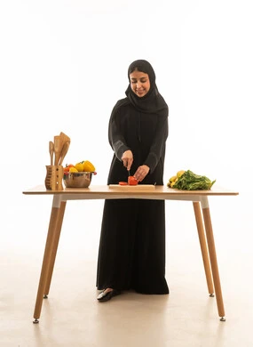 اعداد الطعام في سرور و سعادة , امرأة عربية خليجية سعودية ترتدي عباءة سوداء تقوم بتقطيع الخضروات ,بورتريه خلفية بيضاء الجسم بالطول الكامل