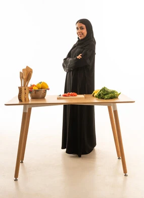 A side view of a smiling Saudi Arabian woman from the Gulf region wearing a black abaya, standing in front of a kitchen table; full-length portrait against a white background.