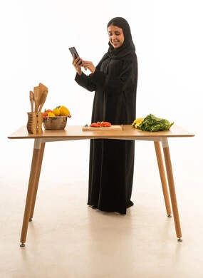 Using a mobile phone to prepare new recipes in happiness; a veiled Saudi Arabian woman from the Gulf wearing a black abaya; full-length portrait with a white background.