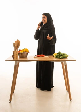 Making a phone call while preparing food; full-length portrait against a white background of a Saudi Arabian Gulf woman wearing a black abaya.