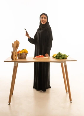 A Saudi Arabian woman wearing a black abaya is holding a mobile phone and looking at something. The portrait is a full-length shot against a white background in front of a food preparation table.