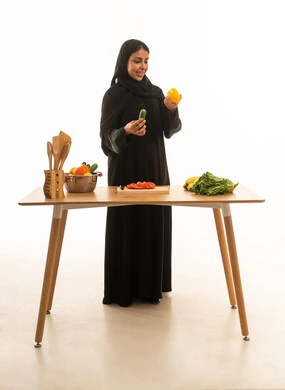 ربة منزل عربية خليجية سعودية ترتدي عباءة سوداء تنظر الى الخضروات الطازجة ,بورتريه خلفية بيضاء, الجسم بالطول الكامل  ,امراة عربية تقف أمام طاولة الطعام