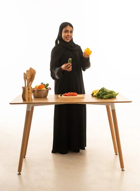 A smiling, veiled Saudi Arabian woman wearing a black abaya is seen carrying fresh vegetables while preparing food. The portrait has a white background and is looking directly at the camera with gestures of happiness. Full-length portrait.