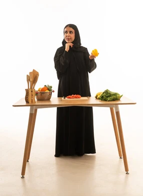 A Saudi Arabian housewife wearing a black abaya, deep in thought, holding vegetables; full-length portrait against a white background, looking upwards.