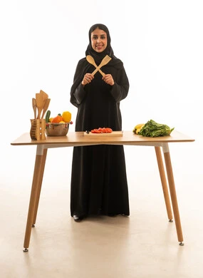 امرأة عربية خليجية سعودية مبتسمة ترتدي عباءة سوداء تحمل ملاعق خشبية مكونة علامة اكس ,بورتريه خلفية بيضاء بالطول الكامل للجسم