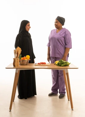 A portrait against a white background of a smiling, veiled Saudi Arabian woman wearing a black abaya, standing with a dark-skinned, foreign African domestic worker wearing a purple uniform, in the kitchen in front of a food preparation table.