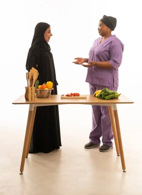 A friendly conversation between a veiled Saudi Arabian woman wearing a black abaya and a dark-skinned foreign domestic worker wearing a purple uniform. (Portrait with a white background, full-length, depicting an Arab woman and her domestic worker at a food preparation table.)