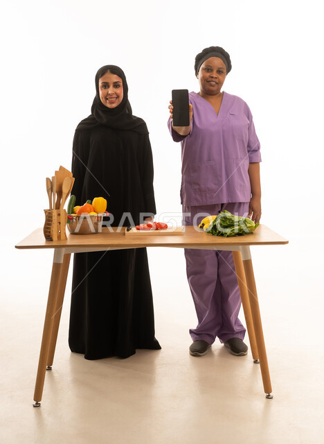 الابتسام للكاميرا مع عرض شاشة هاتف سوداء فارغة ,بورتريه خلفية بيضاء الجسم بالطول الكامل لامرأة عربية خليجية سعودية محجبة ترتدي عباءة سوداء تقف بجانب عاملة منزلية افريقية ملونة داكنة البشرة ترتدي يونيفورم باللون البنفسجي في المطبخ
