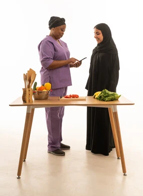 A veiled Saudi Arabian woman wearing a black abaya stands with her dark-skinned African domestic worker, browsing on a smartphone. (This is a full-body portrait against a white background of two women standing at a food preparation table.)