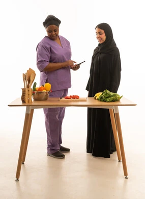سيدة عربية خليجية سعودية محجبة ترتدي عباءة  تتابع عاملتها المنزلية الافريقية سمراء البشرة ترتدي يونيفورم لون بنفسجي اثناء تأدية عملها ,بورتريه خلفية بيضاء للجسم بالطول الكامل لامرأة عربية تقف مع شغالة اجنبية تحمل هاتف محمول أمام طاولة تحضير الطعام