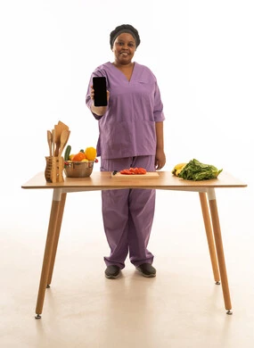 امرأة افريقية سمراء البشرة ترتدي اليونيفورم الخاص للعاملة المنزلية باللون البنفسجي تقف امام طاولة تحضير الطعام بوجه تعتليه ملامح السعادة والابتسام مع عرض شاشة هاتف سوداء فارغة للكاميرا .بورتريه خلفية بيضاء للجسم بالطول الكامل للعاملة مع الخضروات لتحضير طعام صحي .