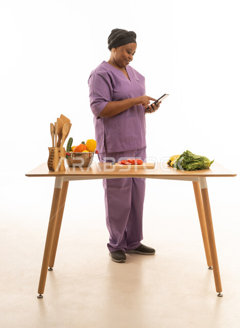 بورترية خلفية بيضاء الجسم بالطول الكامل لعاملة منزل افريقية سمراء البشرة ترتدي يونيفورم باللون البنفسجي تتصفح الهاتف المحمول بايماءات  السعادة والفرح  امام طاولة تحضير الطعام