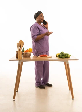 بورتريه خلفية بيضاء لعاملة منزلية سمراء البشرة افريقية ترتدي يونيفورم لون بنفسجي تتحدث بالهاتف المحمول اثناء القيام بتحضير الطعام ,خادمة منزلية تقف امام طاولة تحضير الطعام