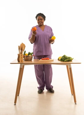 A smiling, dark-skinned African domestic worker wearing a purple uniform and black head covering; a full-body portrait against a white background of a maid holding yellow and orange peppers, standing in front of a food preparation table.