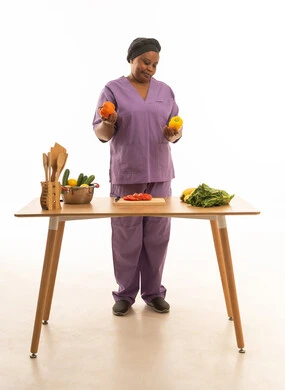 عاملة منزل افريقية سمراء البشرة  ترتدي يونيفورم لون بنفسجي و غطاء راس اسود تمسك فلفل أصفر و برتقالي بايماءات عدم الاعجاب ,بورتريه خلفية بيضاء الجسم بالطول الكامل لخادمة اجنبية ملونة تقف امام طاولة خشبية