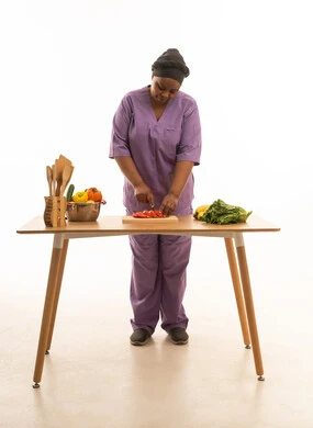 خادمة منزلية ملونة داكنة البشرة ترتدي يونيفورم لون بنفسجي تقوم بتقطيع الخضروات ,بورتريه خلفية بيضاء الجسم بالطول الكامل عاملة منزلية تقف امام طاولة تحضير الطعام