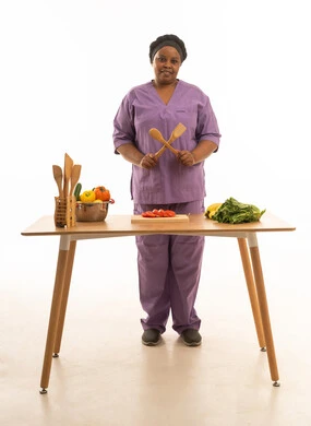 عاملة منزلية سمراء البشرة ترتدي يونيفورم لون بنفسجي تمسك ملاعق خشبية مشيرة بعلامة غلط , بورتريه للجسم بالطول الكامل خلفية بيضاء لشغالة منزلية مبتسمة  تقف امام طاولة تحضير الطعام